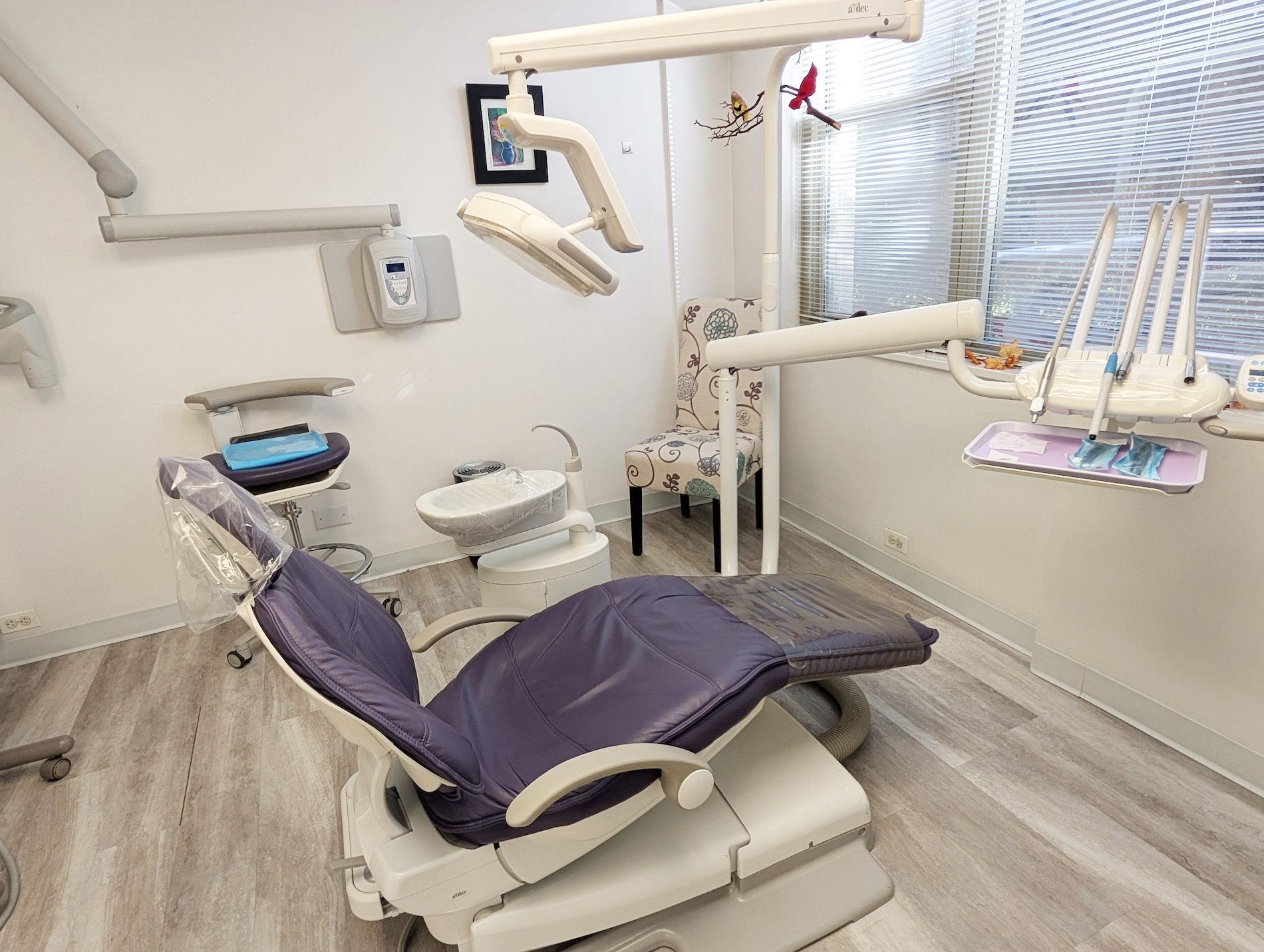 The image shows an interior view of a dental practice with a chair in the foreground, dental equipment, and a clean, well-lit environment.