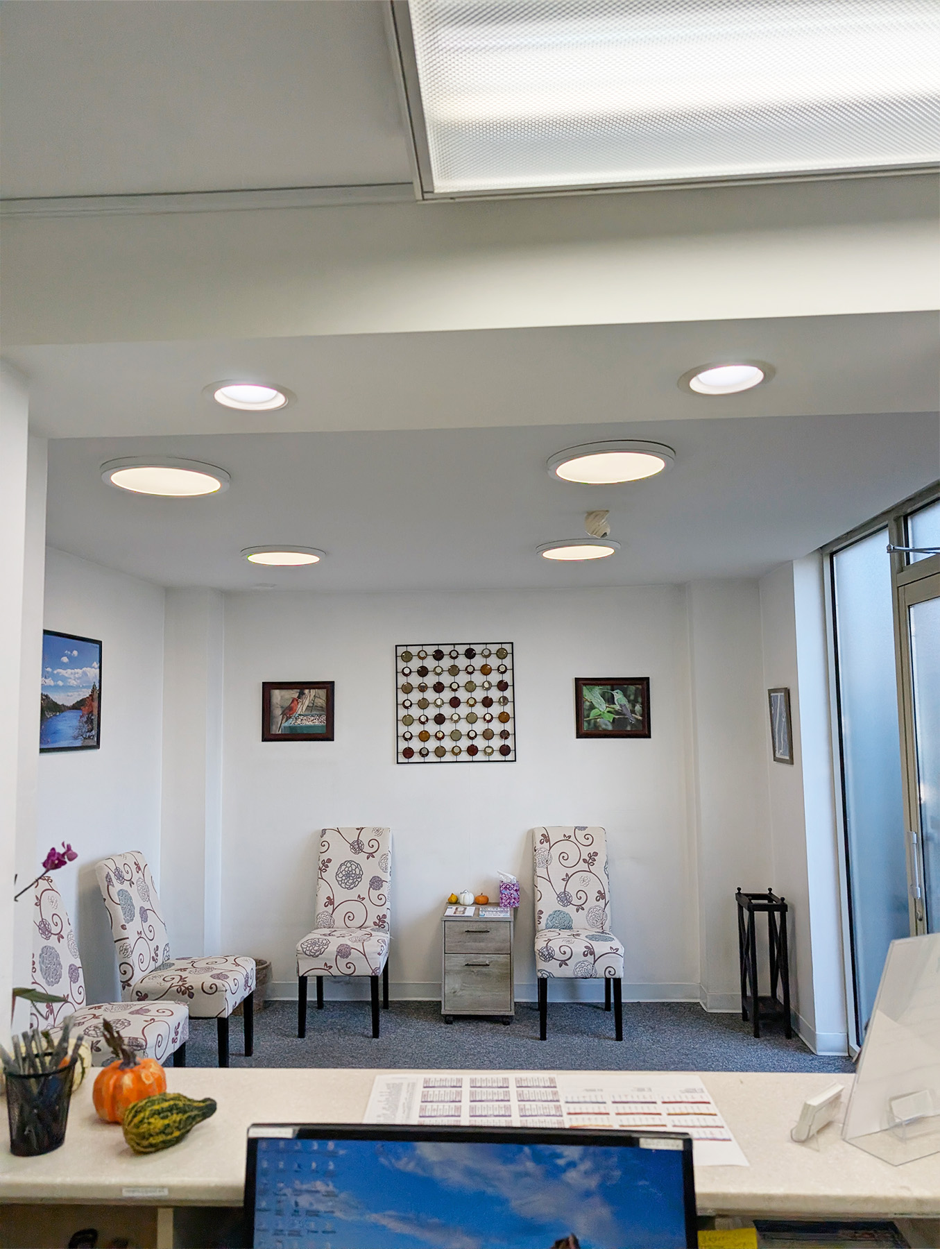 The image shows an interior space with a reception area featuring a patterned wall hanging, a framed photo on the wall, two chairs with floral patterns, a desk with a computer monitor, keyboard, and mouse, and a lamp. Above the desk, there are circular lights affixed to the ceiling.