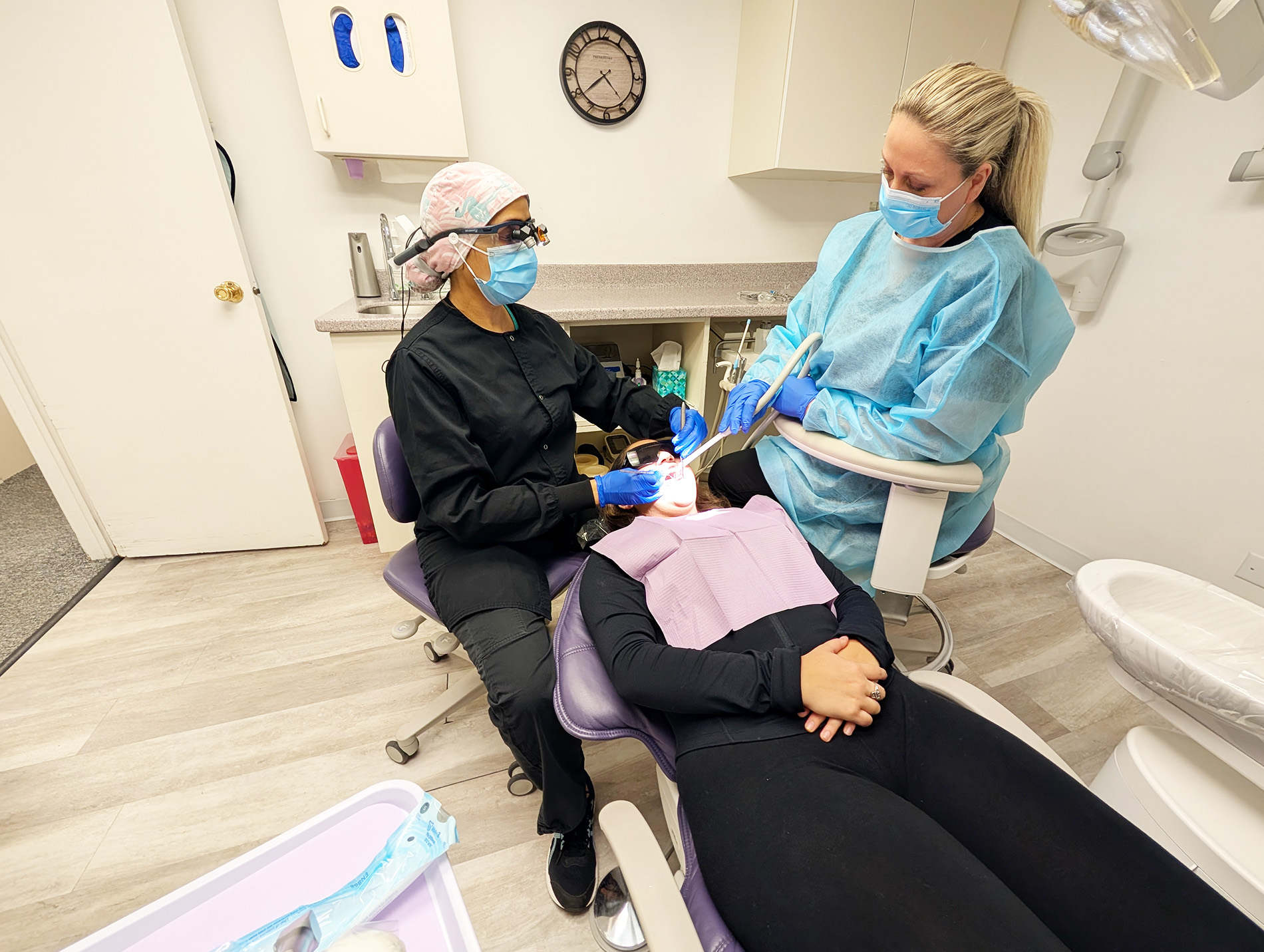 The image shows two individuals wearing medical masks and gloves, attending to a person lying on a dental chair with an open mouth, in a dental office setting.