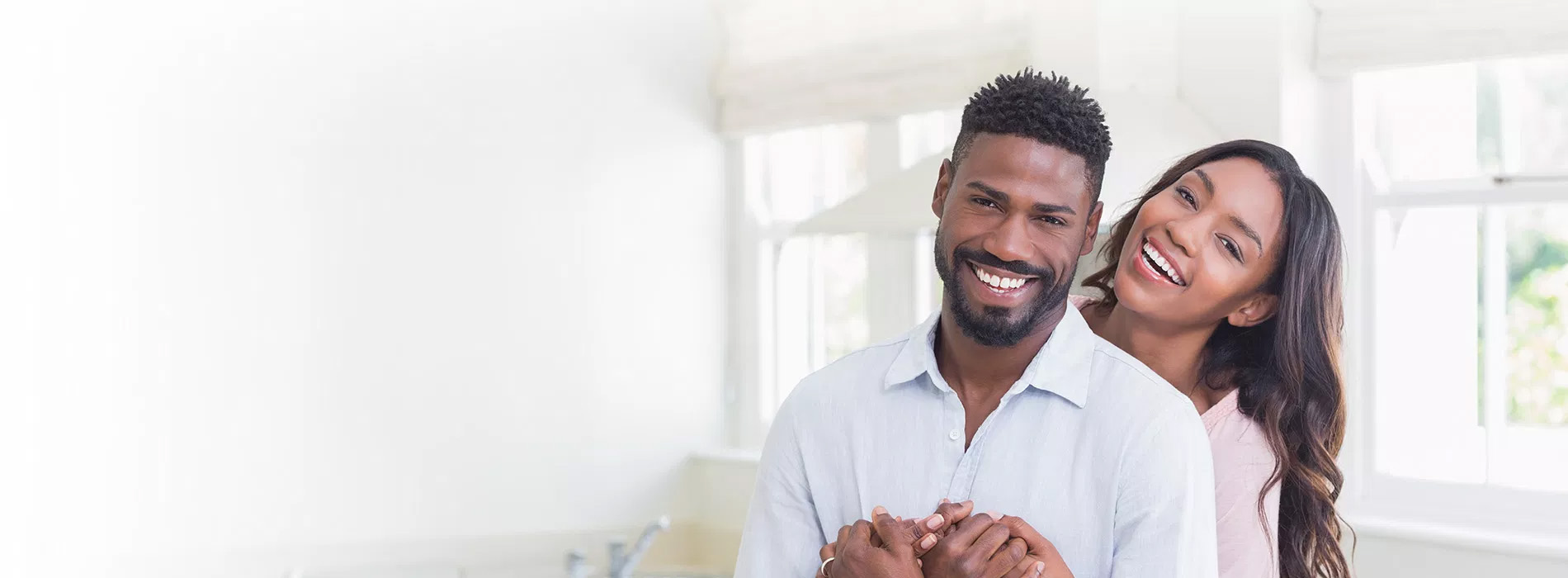 The image shows a man and woman embracing each other with smiles, standing in front of a window.