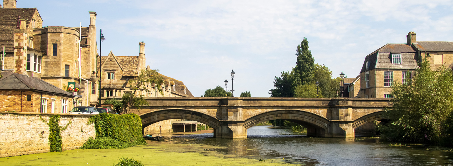 The image displays a serene scene featuring a picturesque village with a stone bridge crossing over a calm river, lined by lush greenery and quaint houses.