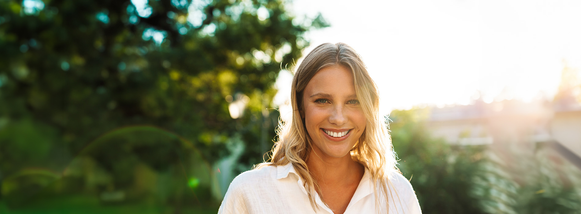 A woman with blonde hair wearing a white top stands outdoors against a blurred background.