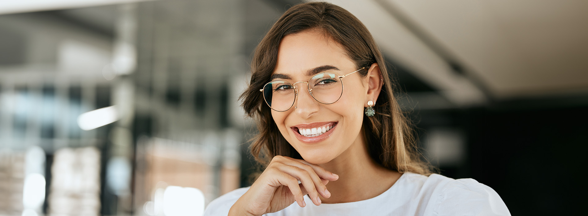 A young woman with glasses smiles at the camera while standing against a blurred background.