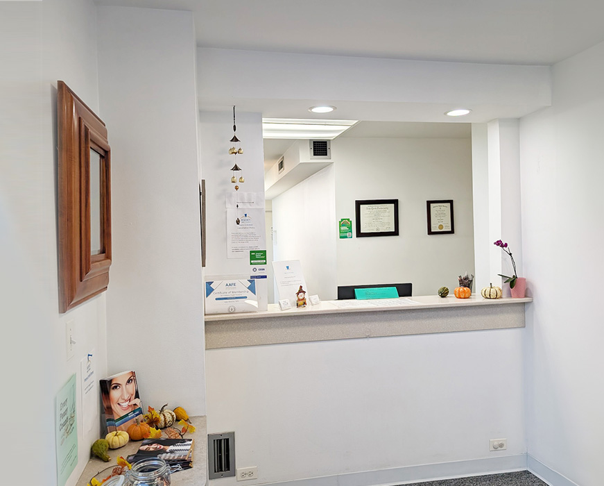 The image shows an interior space with a reception desk, a framed picture on the wall, and various items on the countertop, including a book, a vase with fruit, and what appears to be a sign or notice board.