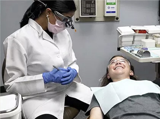 A dental hygienist wearing a face mask and gloves is attending to a patient who is lying down with their eyes closed, receiving dental care in an office setting.