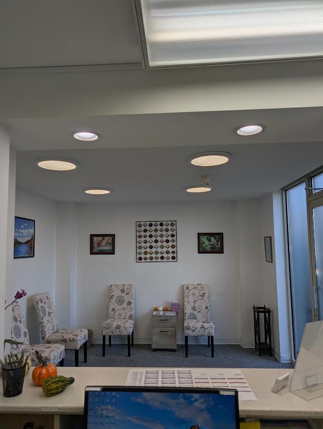 The image shows an interior space, likely a waiting area or reception room, with a white ceiling, recessed lighting, and a decorative wall feature above a seating area that includes two chairs facing each other. There s a framed picture on the wall, a small round table between the chairs, and a sign with text is partially visible at the bottom of the image. The room has a clean, modern appearance.
