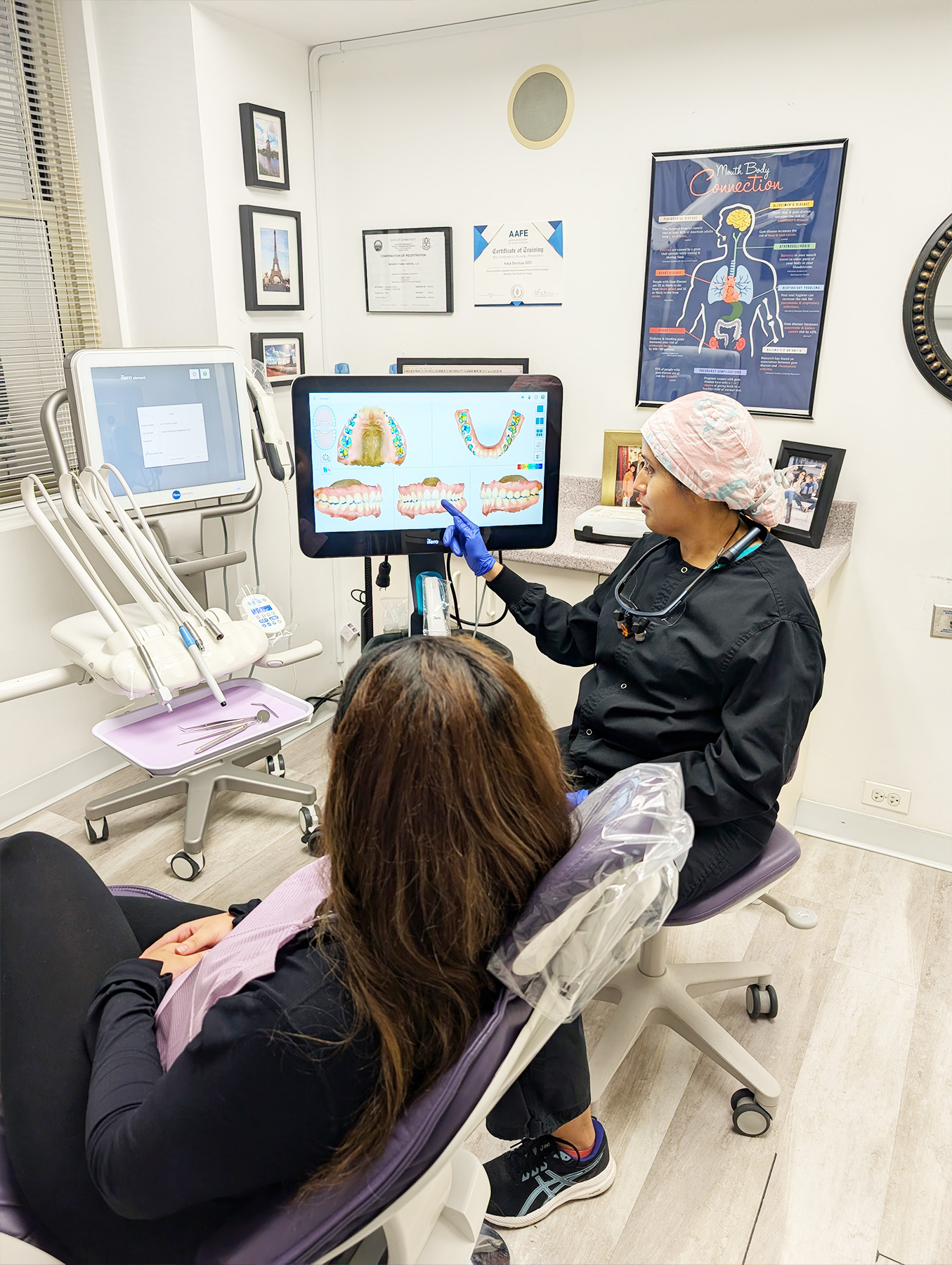 A dental professional in a mask is showing a patient s digital mouth model on a screen while seated at a computer with a monitor, keyboard, and mouse.