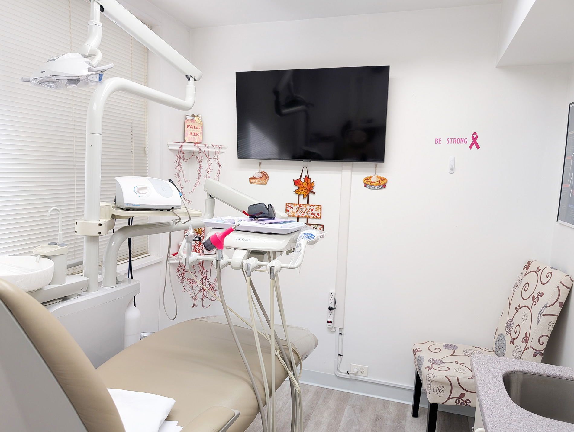 The image depicts a dental office interior with a view of a dental chair and equipment, including a TV screen displaying a heart symbol, a monitor, a stool, and a small decorative tree with a pink ribbon.