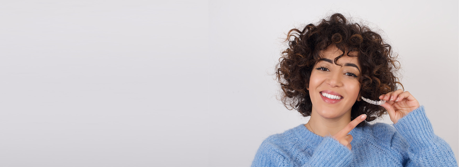 This is a photograph of a person with curly hair smiling at the camera, wearing a blue sweater and holding what appears to be a phone near their ear. The background is plain and light-colored.