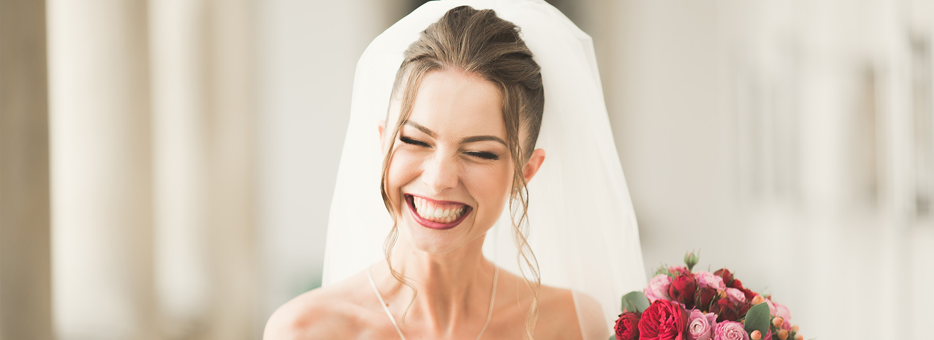 A bride wearing a white veil and holding a bouquet of flowers smiles at the camera during her wedding ceremony.