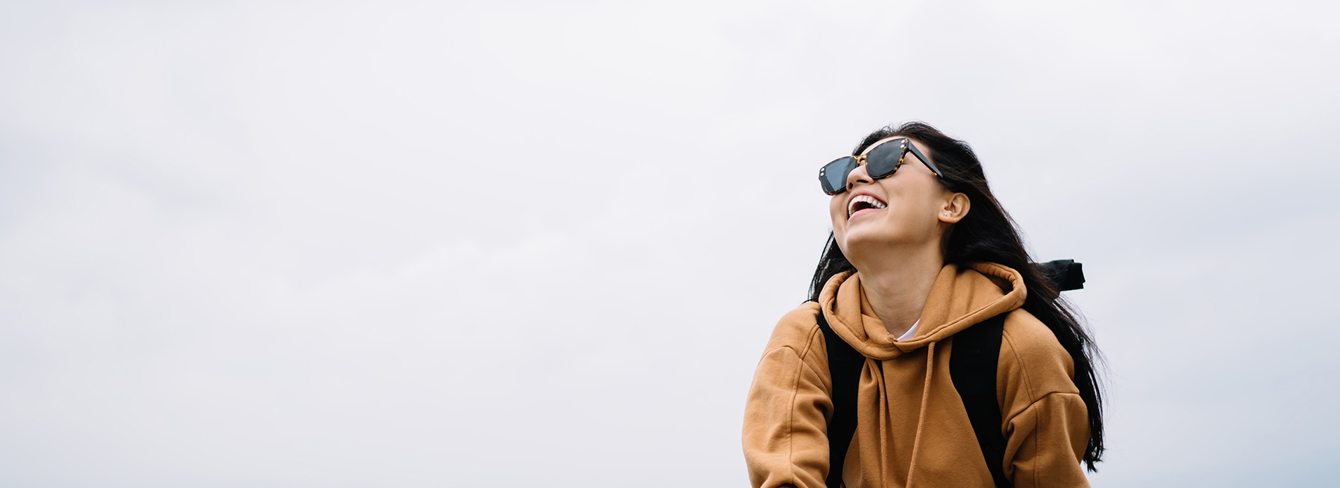 A person with sunglasses stands outdoors, smiling at the camera while wearing a backpack, against an overcast sky.