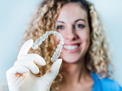 A smiling woman holding up a transparent dental retainer with a clear plastic bridge on top against a white background.