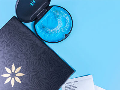 The image shows a dark-colored box with a lid open, revealing a clear container inside holding a blue object that resembles a toothbrush head with bristles. Next to the box, there s a small blue cup and a package of dental floss. The background is plain and light-colored.