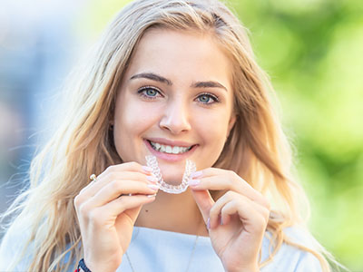 A young woman with blonde hair wearing a white smile brace and holding a clear dental retainer up to her mouth, smiling at the camera.