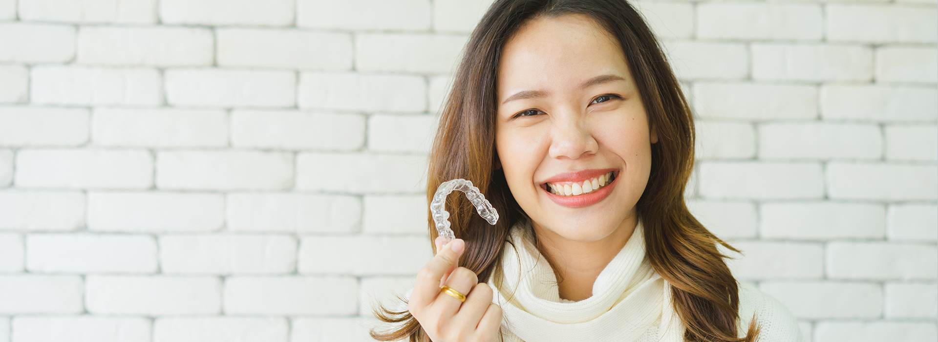 A smiling woman wearing a ring holds up her hand, set against a brick wall background.