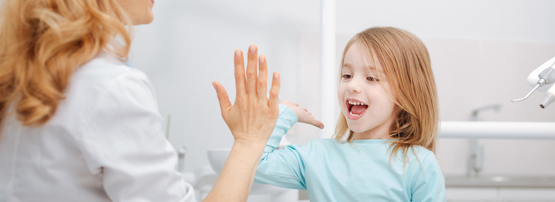 The image shows a woman and a young girl in a dental office setting, with the woman holding out her hand towards the child who appears to be smiling at her.