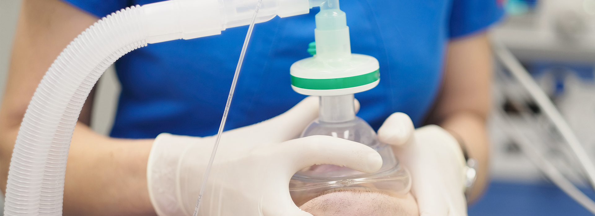 A person wearing gloves and a mask is holding a clear plastic tube with a green cap, which appears to be connected to a medical device, possibly for dispensing medication or collecting samples, set against a background that suggests a medical or laboratory setting.