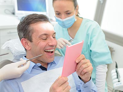 The image shows a man sitting in a dental chair with a large smile, holding a pink card or paper, while looking at it, with a woman standing behind him, wearing a surgical mask and gloves, who appears to be a dental professional.