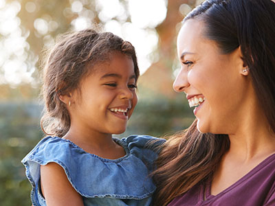 A young girl is smiling at her mother while they stand together outdoors during daylight.