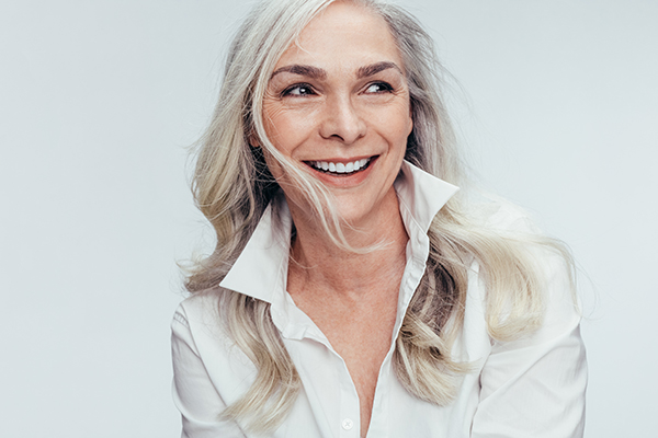 The image shows a woman with short blonde hair smiling at the camera while wearing a white top and a black necklace, against a plain background.