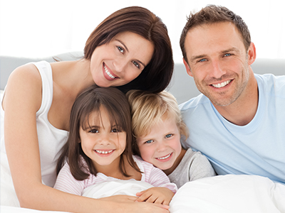 The image shows a family of four people   a man, a woman, and two children   lying together on a bed with smiles on their faces.