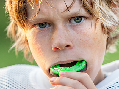 An image of a young male with blonde hair, wearing a football uniform, holding a green sports ball in his mouth while looking directly at the camera.